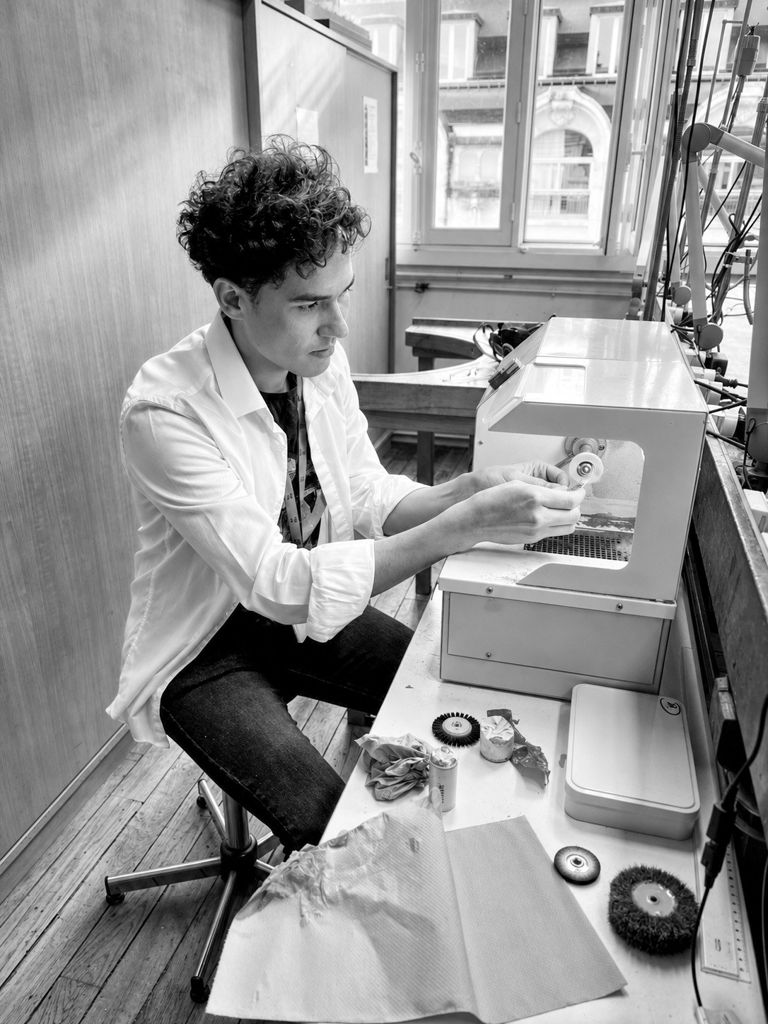 Photographie en noir et blanc de Brendo Delmon assis à un poste de travail de l'atelier, devant une fenêtre.
