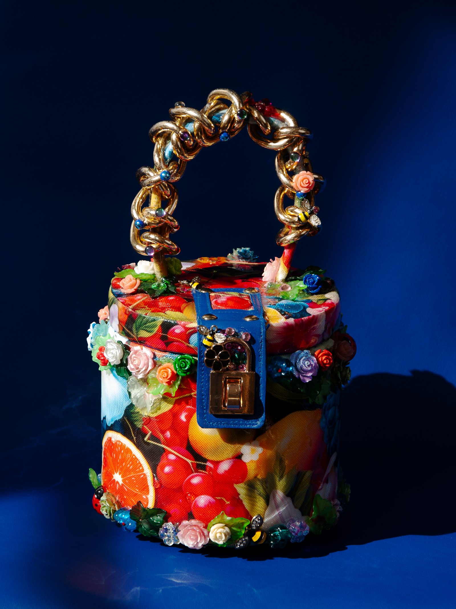 Fiori di Amalfi de Maison Delmon Paris, sac cylindrique à motifs de fruits, fleurs en relief, fermeture bleue et anse à anneaux dorés.